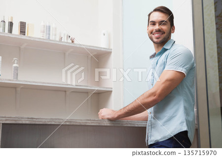 Adult man leaning on rustic counter at salon, wearing blue shirt, shelving with bottles, copy space 135715970