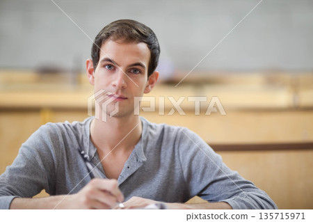 Adult male wearing grey henley sitting at lecture desk, holding pen and writing notes Adult male wearing grey henley sitting at lecture desk, holding pen and writing notes 135715971