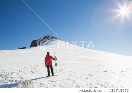 Male skier standing on alpine slope holding skis and pole, wearing red jacket, footprints trailing Male skier standing on alpine slope holding skis and pole, wearing red jacket, footprints trailing 135715972