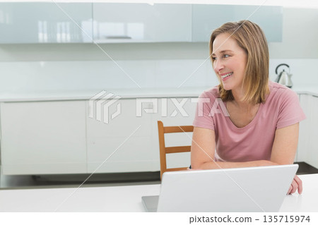 Mature woman wearing pink T-shirt sitting at white kitchen counter using silver laptop, copy space Mature woman wearing pink T-shirt sitting at white kitchen counter using silver laptop, copy space 135715974