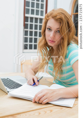 Teenage female studying at wood table with textbook and laptop holding pen wearing turquoise top Teenage female studying at wood table with textbook and laptop holding pen wearing turquoise top 135715975