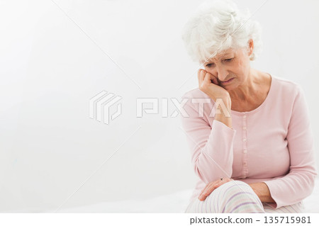Senior female sitting on bed, supporting head, wearing pale pink top, ring visible, copy space Senior female sitting on bed, supporting head, wearing pale pink top, ring visible, copy space 135715981