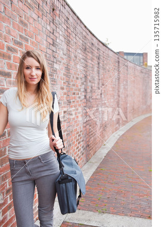 Twenty-something woman in white top jeans posing by red brick wall with messenger bag, copy space 135715982