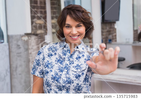 Female hairstylist smiling and holding scissors while standing in salon wearing blue floral blouse 135715983