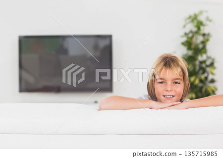 Boy child leaning on sofa wearing T-shirt at home with wall TV and plant, copy space 135715985