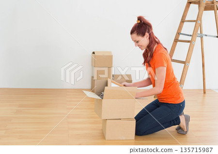 In room female in orange-tee kneeling unpacking framed item from moving box near ladder, copy space 135715987