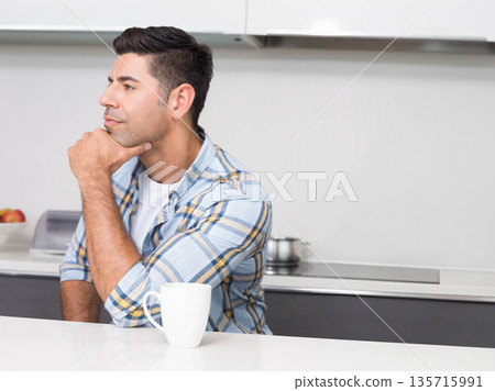 Man sitting at white kitchen countertop wearing plaid shirt, resting chin, holding mug 135715991