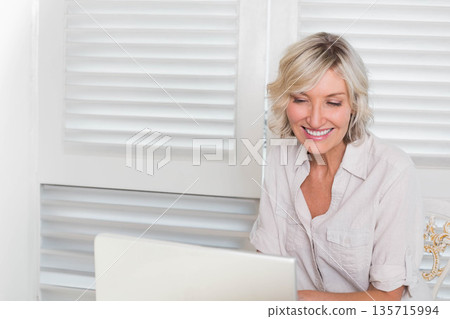 Senior woman sitting at desk in shirt sleeves rolled smiling at laptop with shutters, copy space Senior woman sitting at desk in shirt sleeves rolled smiling at laptop with shutters, copy space 135715994