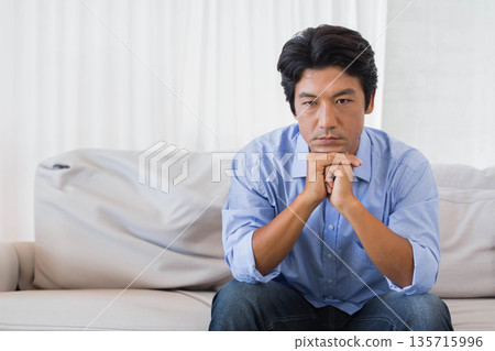 Asian mid-adult man sitting on sofa in living room with white curtains wearing blue shirt, jeans 135715996