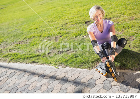 Senior woman sitting at park in lavender fixing skates with yellow wheels and pads, copy space Senior woman sitting at park in lavender fixing skates with yellow wheels and pads, copy space 135715997