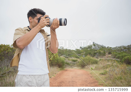 Man in his twenties standing on reddish dirt trail holding DSLR with long telephoto lens Man in his twenties standing on reddish dirt trail holding DSLR with long telephoto lens 135715999