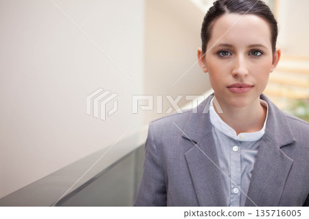 Businesswoman standing by glass railing in office lobby wearing grey blazer and blouse, copy space 135716005