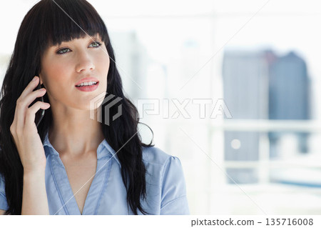 Adult woman holding phone to ear, looking upward near office window in collared blouse, copy space 135716008
