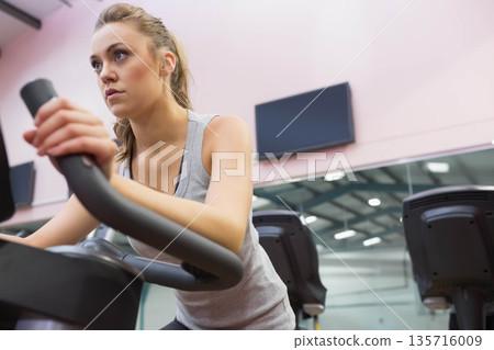 Woman in gray sleeveless top gripping handlebars and pedaling on cardio bike in gym, mirror visible Woman in gray sleeveless top gripping handlebars and pedaling on cardio bike in gym, mirror visible 135716009