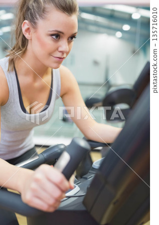 Female exercising on elliptical at gym, gripping handles and watching console, wearing grey tanktop Female exercising on elliptical at gym, gripping handles and watching console, wearing grey tanktop 135716010