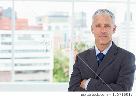 Senior man in dark gray suit, navy tie standing by floor-to-ceiling window at office, copy space Senior man in dark gray suit, navy tie standing by floor-to-ceiling window at office, copy space 135716012