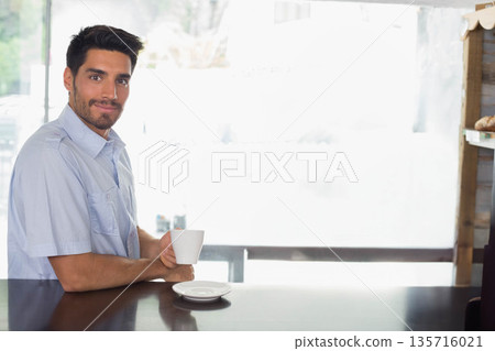 Man in blue shirt sitting at wooden cafe counter holding white mug with saucer, copy space 135716021