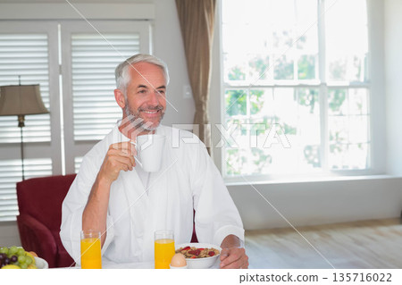 Senior man sitting in white robe at dining table near window, eating cereal, holding orange juice 135716022