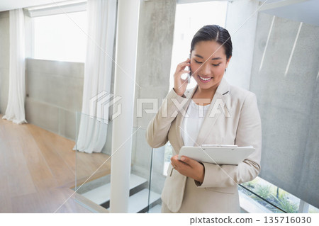 Asian woman standing, talking on phone and holding clipboard in beige suit on mezzanine, copy space 135716030