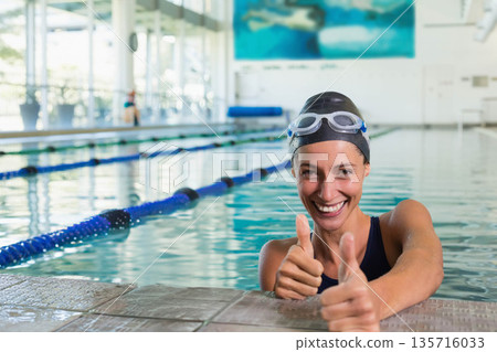 Adult female swimmer leaning on lane pool edge holding coping and giving thumbs up, copy space 135716033