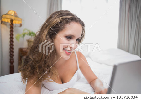 Woman reclining on bed wearing white camisole, smiling while using laptop in bedroom 135716034