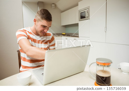 Male sitting at kitchen table working on silver laptop in orange striped t-shirt with French press Male sitting at kitchen table working on silver laptop in orange striped t-shirt with French press 135716039