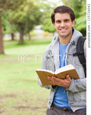 Man holding open hardcover book at chest in park, wearing denim jacket, backpack strap, copy space 135716042