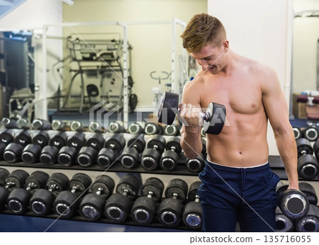 Shirtless man mid-20s doing biceps curl wearing navy shorts by dumbbell rack and mirror, copy space 135716055