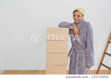 African American woman wearing robe, headscarf, leaning on boxes in room near ladder, copy space African American woman wearing robe, headscarf, leaning on boxes in room near ladder, copy space 135716058