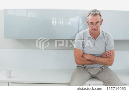 Man sitting on white counter in kitchen with white mug, crossing arms, wearing glasses, copy space Man sitting on white counter in kitchen with white mug, crossing arms, wearing glasses, copy space 135716063