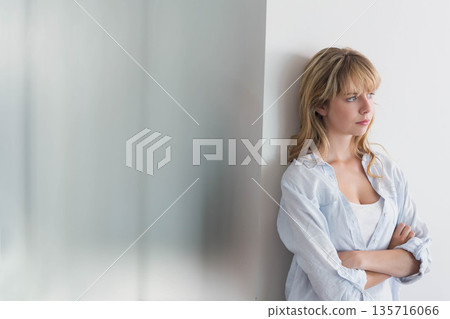 Woman mid-20s leaning on white wall by frosted glass, wearing shirt, looking right, copy space 135716066