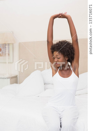 African American woman sitting on bed stretching arms overhead in bedroom wearing white sleepwear African American woman sitting on bed stretching arms overhead in bedroom wearing white sleepwear 135716071