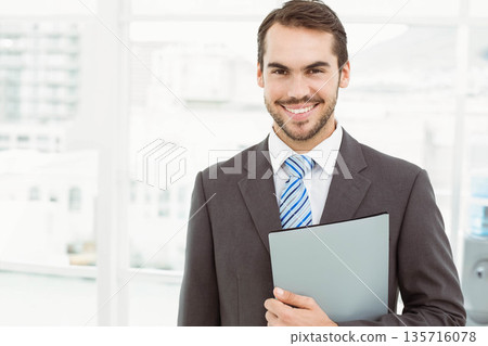 Male professional in dark gray suit standing holding light-gray binder by office window, copy space Male professional in dark gray suit standing holding light-gray binder by office window, copy space 135716078