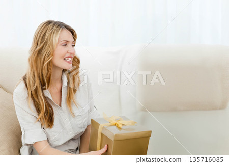 Blonde woman sitting on beige sofa holding gold gift box wearing light button-up shirt, copy space Blonde woman sitting on beige sofa holding gold gift box wearing light button-up shirt, copy space 135716085