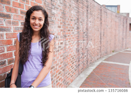 Woman in mid-20s standing, leaning on red brick wall by curved walkway, backpack strap, copy space Woman in mid-20s standing, leaning on red brick wall by curved walkway, backpack strap, copy space 135716086