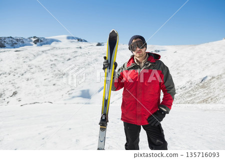 Adult male standing snowy slope holding yellow skis, wearing red jacket and goggles, copy space Adult male standing snowy slope holding yellow skis, wearing red jacket and goggles, copy space 135716093