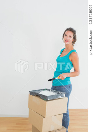 Woman in turquoise top holding roller, tray on moving boxes, painting wall at home, copy space 135716095