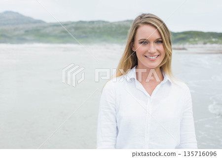 Woman in late twenties standing on shoreline at quiet beach, smiling and wearing white button shirt Woman in late twenties standing on shoreline at quiet beach, smiling and wearing white button shirt 135716096