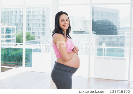 Pregnant woman standing by tall windows in bright studio cradling belly in maternity leggings Pregnant woman standing by tall windows in bright studio cradling belly in maternity leggings 135716100
