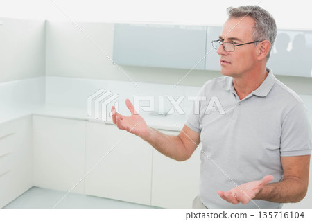 Senior man wearing gray polo and glasses gesturing in white kitchen, phone on counter, copy space Senior man wearing gray polo and glasses gesturing in white kitchen, phone on counter, copy space 135716104