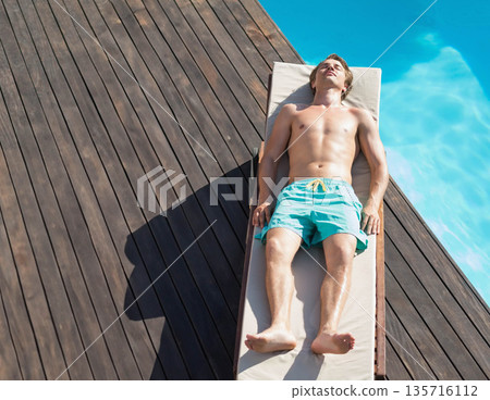 Man reclining on lounge chair on wooden deck beside turquoise pool wearing teal trunks, copy space Man reclining on lounge chair on wooden deck beside turquoise pool wearing teal trunks, copy space 135716112