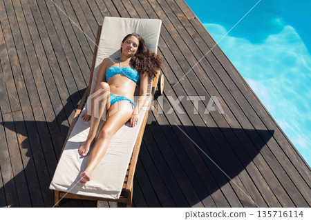 Woman in her twenties sunbathing on wooden chaise on deck by turquoise pool in blue bikini Woman in her twenties sunbathing on wooden chaise on deck by turquoise pool in blue bikini 135716114