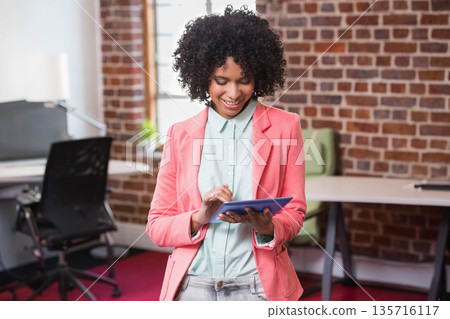 Woman in coral blazer holding and tapping blue tablet while standing in modern office Woman in coral blazer holding and tapping blue tablet while standing in modern office 135716117