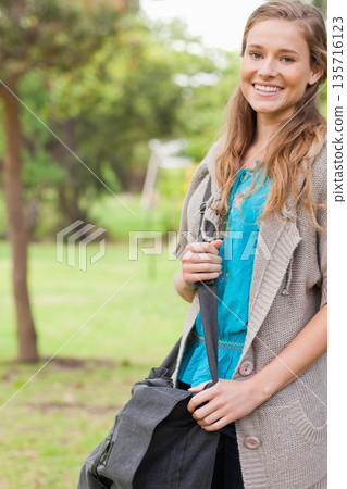 Female standing at park holding gray bag, smiling, wearing turquoise blouse and knit cardigan 135716123