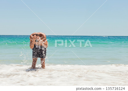 Man bending, splashing seawater onto face in shallow foamy surf at sandy beach wearing dark trunks Man bending, splashing seawater onto face in shallow foamy surf at sandy beach wearing dark trunks 135716124