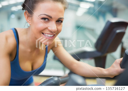 Female gym-goer smiling, leaning and holding cardio machine handles wearing blue sports tank at gym Female gym-goer smiling, leaning and holding cardio machine handles wearing blue sports tank at gym 135716127