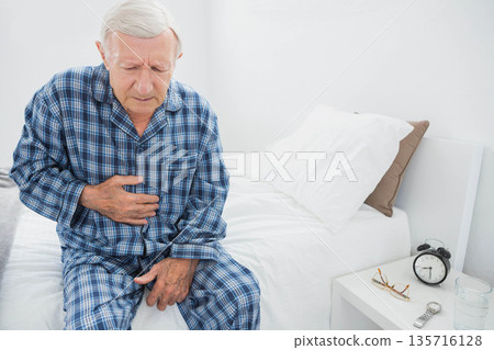 Senior man sitting on bed edge clutching stomach wearing blue plaid pajamas with clock, copy space Senior man sitting on bed edge clutching stomach wearing blue plaid pajamas with clock, copy space 135716128