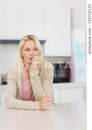 Mid adult female sitting at light countertop, wearing beige cardigan, touching mouth, looking left 135716134