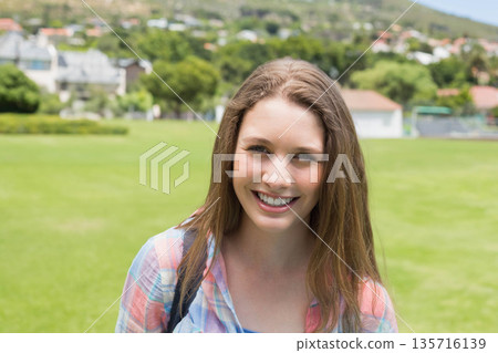 Teenage female standing smiling facing camera on grassy field in pastel plaid top backpack strap Teenage female standing smiling facing camera on grassy field in pastel plaid top backpack strap 135716139