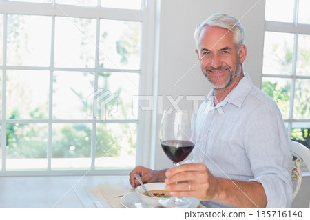 Senior male blue-shirt sitting at dining table grid-window holding wine glass and bowl, copy space Senior male blue-shirt sitting at dining table grid-window holding wine glass and bowl, copy space 135716140